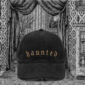 Könnte beinhalten: Schwarze Cord-Baseballkappe mit dem Wort "haunted" in goldfarbenem Faden gestickt. Die Kappe hat einen gebogenen Schirm und eine runde Krone. Der Hintergrund zeigt eine Schwarz-Weiß-Illustration eines Raumes.