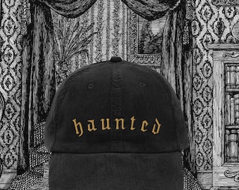 May include: Black corduroy baseball cap with the word "haunted" embroidered in gold thread. The cap has a curved brim and a rounded crown. The background features a black and white illustration of a room.