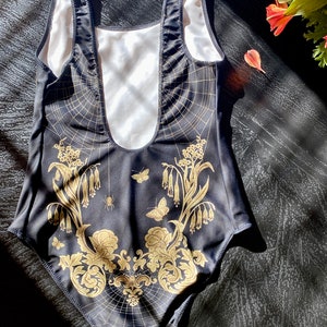 May include: Black swimsuit with a gold floral and spiderweb design. The swimsuit has a scoop neck and a low back.