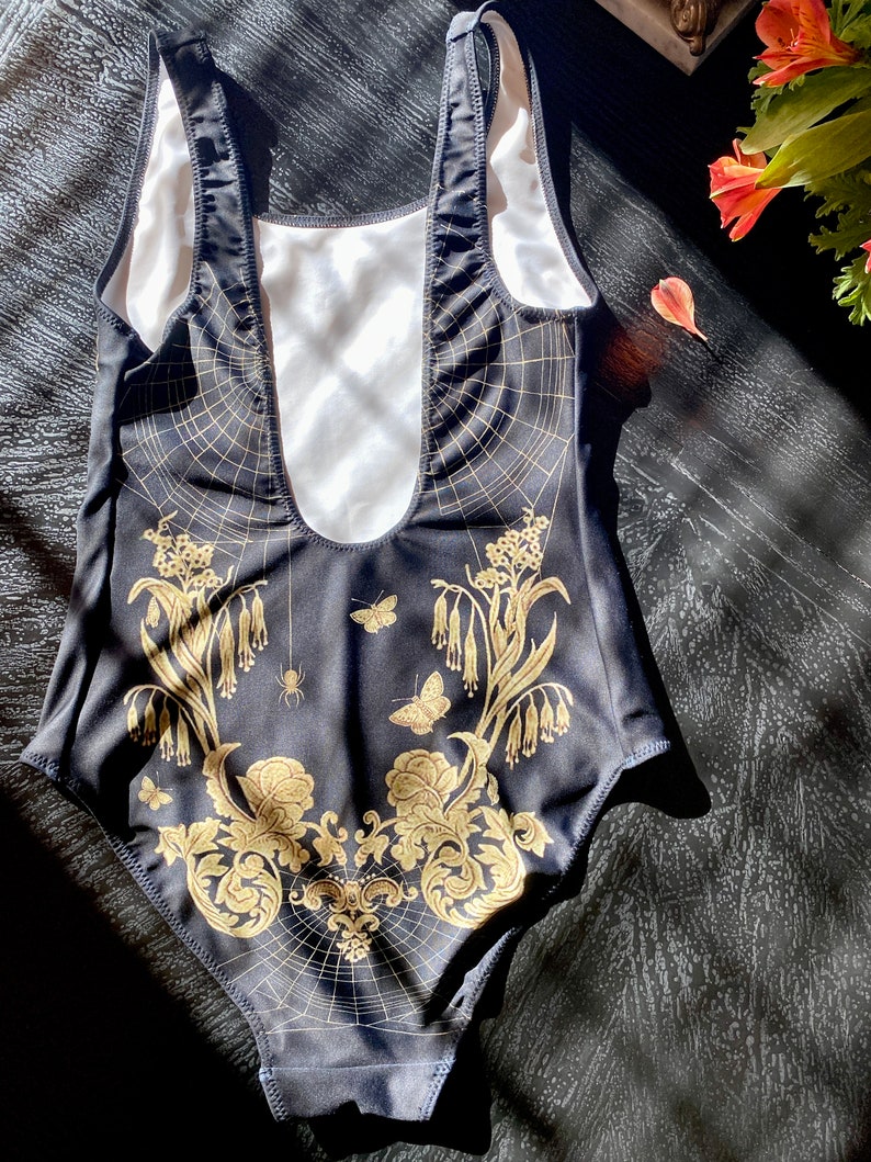 May include: Black swimsuit with a gold floral and spiderweb design. The swimsuit has a scoop neck and a low back.