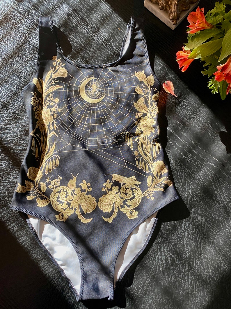 May include: Black swimsuit with a gold spiderweb design featuring a crescent moon and a spider. The design also includes floral and leaf motifs.