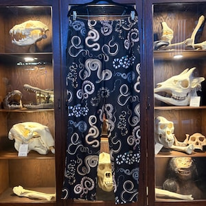 May include: Black trousers featuring a repeating pattern of coiled snakes in shades of white, grey, and brown. The trousers have a straight-leg cut, a waistband with belt loops, and are displayed on a black hanger against a wooden cabinet.