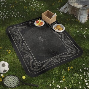 May include: Black and white picnic blanket featuring a spiderweb design and a border of snakes. The blanket is laid out on green grass with a picnic basket, fruit, and snacks.