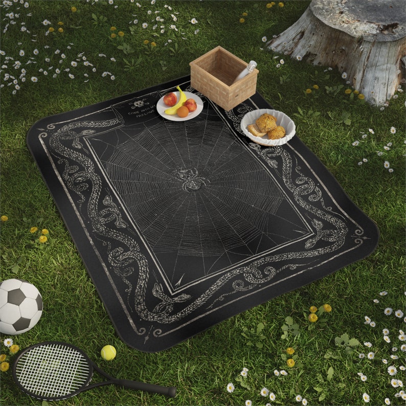 May include: Black and white picnic blanket featuring a spiderweb design and a border of snakes. The blanket is laid out on green grass with a picnic basket, fruit, and snacks.