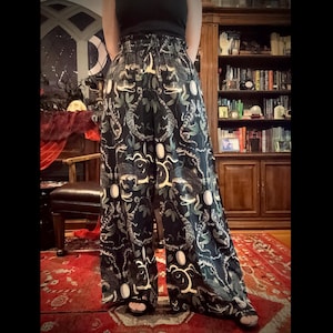 May include: Black palazzo trousers featuring a repeating pattern of snakes, leaves, and botanical elements in green, white, and yellow. The trousers have a wide leg and an elasticated waistband.