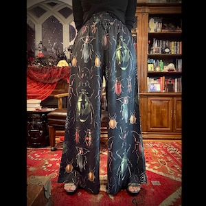 May include: Black palazzo pants with a repeating pattern of colorful beetles. The insects are in shades of green, gold, red, and brown. The pants have a wide leg and an elastic waistband. The background is a room with a red rug and a wooden bookcase.