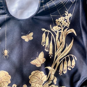 May include: Black swimsuit with a gold spiderweb, floral, and butterfly design.