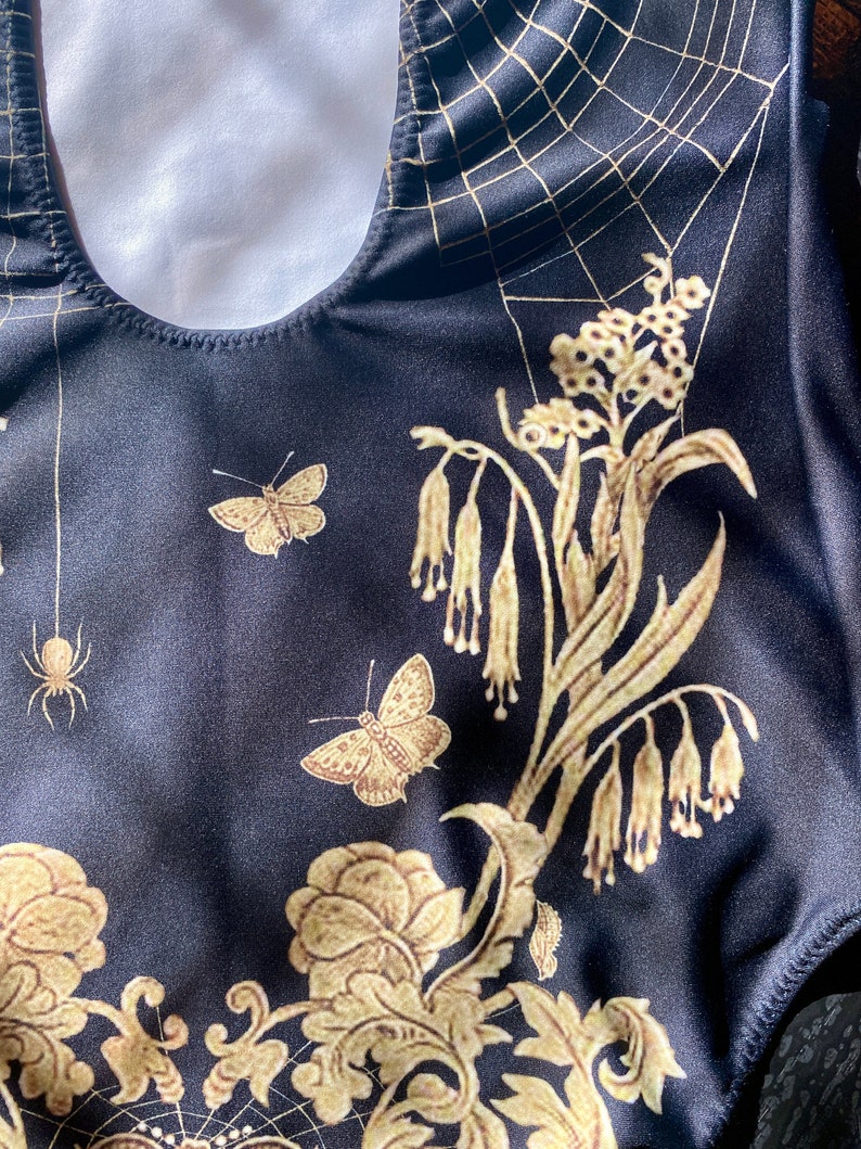 May include: Black swimsuit with a gold spiderweb, floral, and butterfly design.