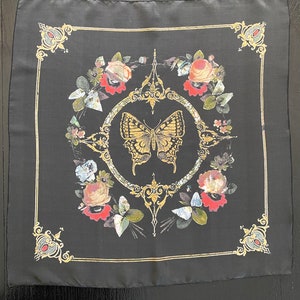 May include: Black silk scarf with a gold butterfly and floral design. The butterfly is in the center of the scarf and is surrounded by flowers and leaves. The scarf has a gold border.