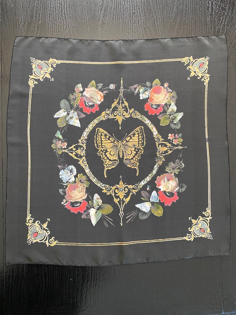 May include: Black silk scarf with a gold butterfly and floral design. The butterfly is in the center of the scarf and is surrounded by flowers and leaves. The scarf has a gold border.