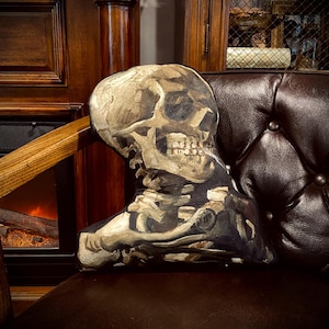 May include: A black leather armchair with a skull-shaped pillow. The pillow is printed with a detailed image of a human skull and skeleton.