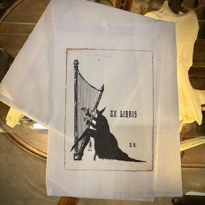 May include: A white rectangular textile featuring a black ink illustration of a figure in a long coat playing a harp. The design includes the text "EX LIBRIS" and the initials "E.B." with a small date stamp of 1916 near the base of the harp.