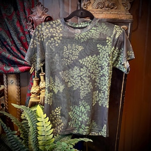 May include: A short-sleeved crew neck t-shirt made of a semi-sheer dark gray mesh fabric. The garment features an all-over print of small, detailed fern leaves in shades of light green and yellow, distributed across the front and sleeves.