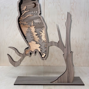 May include: A wooden owl sculpture with a detailed cutout design of a forest scene inside. The owl is perched on a branch and has a base.