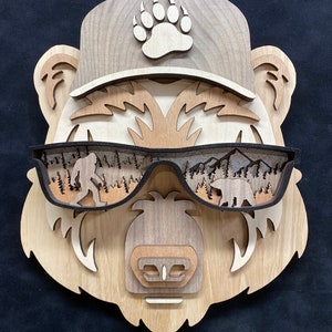 May include: Wooden bear head wall art with a brown baseball cap featuring a paw print design and sunglasses with a forest scene. The bear has a brown nose and a brown mouth.