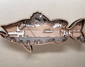 May include: A wooden wall art sculpture of a fish with a lake scene inside. The scene includes trees, mountains, a boat, and people fishing.