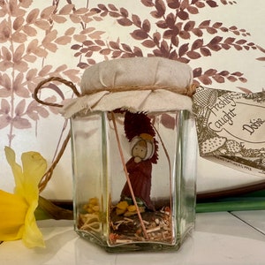 May include: A glass jar with a fabric lid and a brown twine tie. Inside the jar is a small figurine of a person wearing a red robe. The jar has a label that reads "Freshly Caught Dobby" and "Friend Catchers 1912".