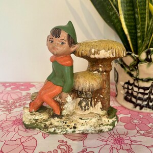 May include: A green and orange elf figurine sitting on a mushroom-shaped base. The elf has a pointed hat and large ears. The mushroom is painted in shades of brown and yellow.