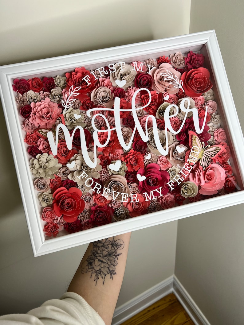 Pu&ograve; includere: Una scatola di ombra incorniciata di bianco piena di fiori di carta rosa, rossi e crema. Le parole "First My Mother Forever My Friend" sono scritte in lettere bianche sui fiori.