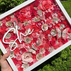 Pu&ograve; includere: Una scatola a ombre incorniciata di bianco piena di fiori di carta rosa e rossi e farfalle. Il testo "She motivates, inspires, empowers, she is me, I am she." &egrave; scritto in bianco sulla parte anteriore della scatola.