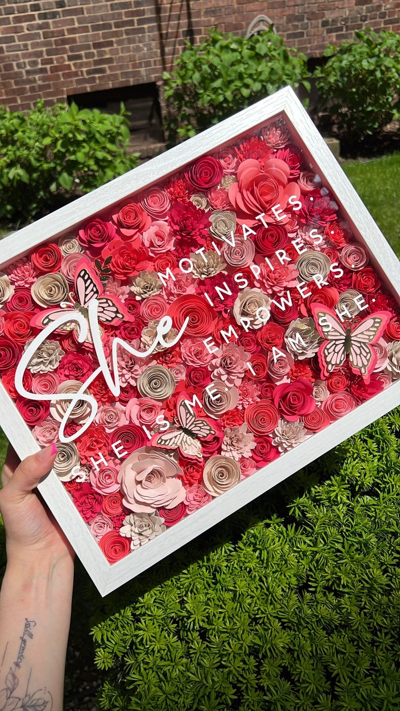 Pu&ograve; includere: Una scatola a ombre incorniciata di bianco piena di fiori di carta rosa e rossi e farfalle. Il testo "She motivates, inspires, empowers, she is me, I am she." &egrave; scritto in bianco sulla parte anteriore della scatola.