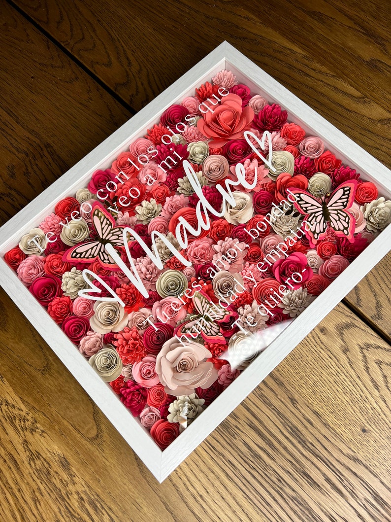 Pu&ograve; includere: Una cornice bianca piena di fiori di carta rosa, rossi e crema. I fiori sono disposti a forma di cuore con la parola "Madre" in lettere bianche al centro. Il testo "Te veo con los ojos que veo a mi Madre: amor, bondad, humildad y empatia. Te quiero." &egrave; scritto in lettere bianche intorno ai fiori.