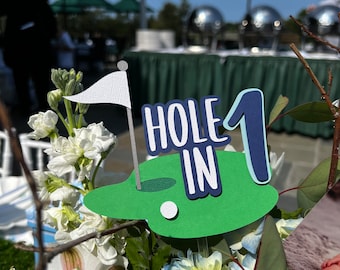 Op de afbeelding: Een groene en blauwe papieren uitsnijding van een golfgreen met een witte golfbal en een vlag. De uitsnijding zegt "Hole in 1" in witte en blauwe letters. De uitsnijding is geplaatst in een boeket witte en blauwe bloemen.