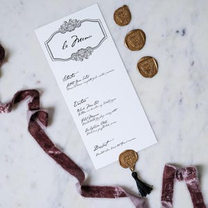 May include: A white menu card with the title "le Menu" in elegant script, accompanied by three gold wax seals and a burgundy velvet ribbon. The menu lists starters, entrees, and dessert.