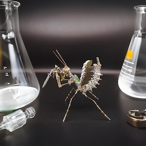 May include: A detailed steampunk-style metal praying mantis sculpture with a small knife. The insect has intricate mechanical details, including gears and wires, with a mix of silver and gold tones. Two glass beakers are in the background.