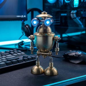 May include: A small, metallic robot figurine constructed from brass-toned components. It features a rounded body, two articulated arms, and two dome-shaped feet. The head has two prominent circular eyes that emit a bright, glowing blue light.