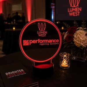 May include: A red illuminated sign with the text "LUMEN WEST performance lighting systems" in a circular frame. The sign sits on a black base. A stack of brochures with the text "BRIGHTER TOGETHER" is in the foreground.