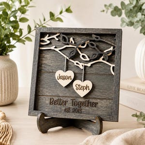 K&ouml;nnte beinhalten: Ein quadratisches, dunkelgraues Holzschild mit einem geschichteten Astmotiv und zwei h&auml;ngenden, hellen Holzherzen, in die die Namen Jason und Steph eingraviert sind. Das Schild enth&auml;lt den gravierten Text Better Together und est 2013.