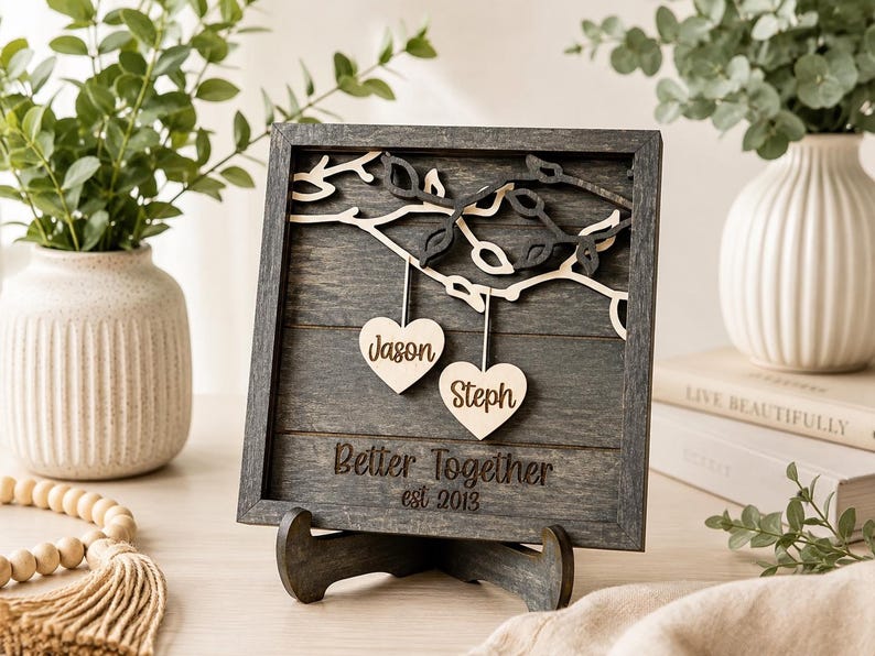 K&ouml;nnte beinhalten: Ein quadratisches, dunkelgraues Holzschild mit einem geschichteten Astmotiv und zwei h&auml;ngenden, hellen Holzherzen, in die die Namen Jason und Steph eingraviert sind. Das Schild enth&auml;lt den gravierten Text Better Together und est 2013.