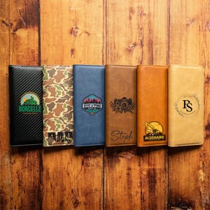 May include: Six rectangular wallets arranged in a row. The first is black carbon fiber with a Borcelle logo. The second has a camouflage pattern with the word DAD. The third is blue with a Dye & Pine logo. The fourth is brown with a camera graphic and the name Steph. The fifth is tan with an Aldenaire construction logo. The sixth is light tan with a circular floral monogram featuring the initials PS and the date 09-21-2027.