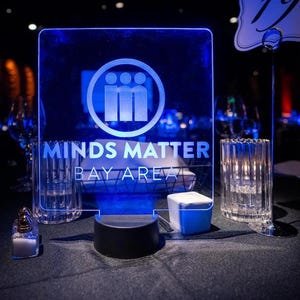 May include: A glowing blue acrylic sign with the words "MINDS MATTER BAY AREA" and a circular logo featuring three figures. The sign is illuminated, creating a striking visual effect, and is set on a table with other decorative elements.