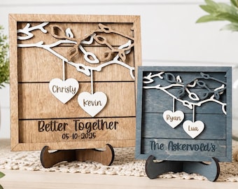 Könnte beinhalten: Zwei quadratische Holzschilder auf Ständern. Das größere Schild ist hellbraun mit weißen Ästen, zwei weißen Herzen mit den Namen Christy und Kevin sowie dem Text Better Together 05-10-2025. Das kleinere Schild ist blau mit weißen Ästen, zwei weißen Herzen mit den Namen Ryan und Lua sowie dem Text The Askervold's.