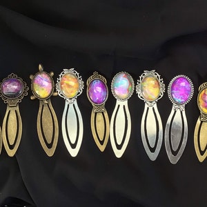 May include: A set of ten colorful bookmarks with different designs. Each bookmark features a round, glass-like charm with a unique abstract pattern in shades of pink, purple, yellow, and blue. The charms are set in ornate metal frames with a vintage aesthetic. The bookmarks are made of metal and have a gold or silver finish.