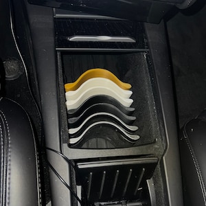 May include: A stack of multi-colored car coasters in a vehicle's center console. The coasters are in shades of yellow, white, gray, and black. The console is black and has a storage compartment.