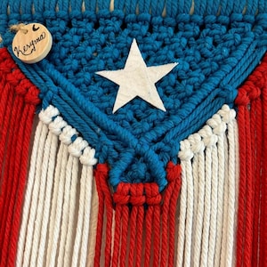 Puede incluir: Un tapiz de macramé azul, blanco y rojo con una estrella blanca en el centro. El tapiz tiene una etiqueta de madera con la palabra "Keruma" escrita en ella.