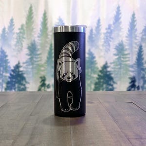 May include: Black insulated tumbler featuring a detailed etched design of a red panda. The stainless steel tumbler has a silver lid and a slender, cylindrical shape. The red panda illustration is in a walking pose, with a fluffy tail and detailed fur.
