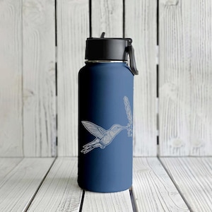 May include: A navy blue water bottle with a black lid and handle. A white hummingbird design is etched on the side, depicting the bird in flight near a flower. The bottle is set against a white wooden background.