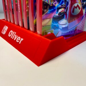 May include: Red plastic Nintendo Switch game case holder with the name "Oliver" printed on the front. The holder has slots for multiple game cases and a raised section for a single case.