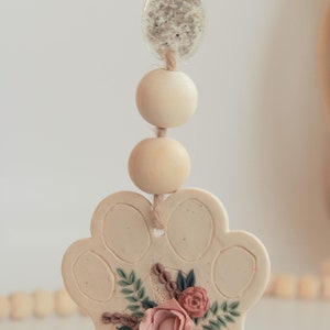 May include: A beige ceramic paw print charm with a floral design in shades of pink, green, and brown. The charm is attached to a string of wooden beads and a clear glass bead with a speckled design.