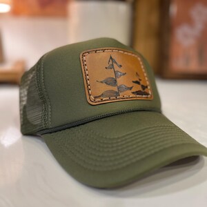 May include: Olive green trucker hat with a brown leather patch featuring a tree design. The hat has a mesh back and a curved brim. The patch is stitched onto the hat with a light brown thread.
