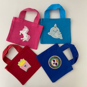 May include: Four small tote bags with different colorful designs. The bags are made of a canvas material and have a single handle. The designs include a unicorn, a narwhal, french fries, and a sloth.
