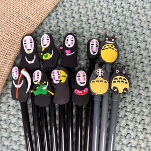 May include: A set of 12 black pens with various colorful cartoon character toppers, including a black and white character with a scythe, a character with a pink heart, a yellow character with a red mouth, and a blue character with a red mouth.