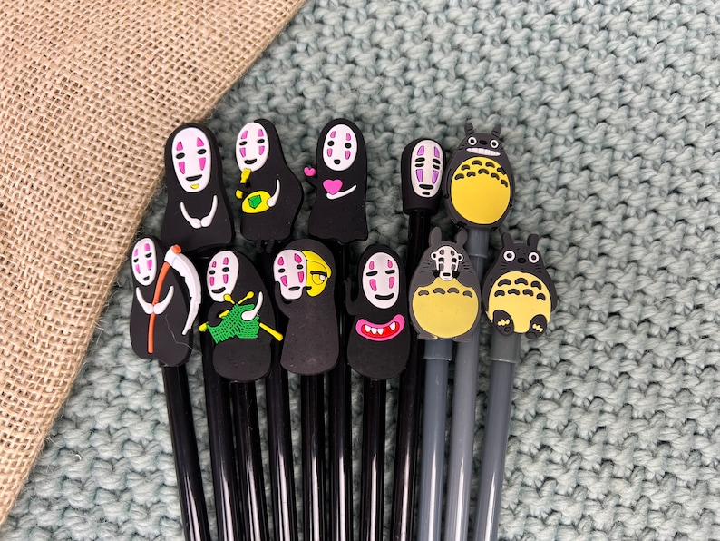 May include: A set of 12 black pens with various colorful cartoon character toppers, including a black and white character with a scythe, a character with a pink heart, a yellow character with a red mouth, and a blue character with a red mouth.