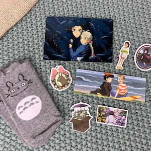 May include: A collection of Studio Ghibli themed items, including a pair of grey socks with a Totoro design, a postcard featuring Howl and Sophie from Howl's Moving Castle, and several stickers featuring characters from different Ghibli films.