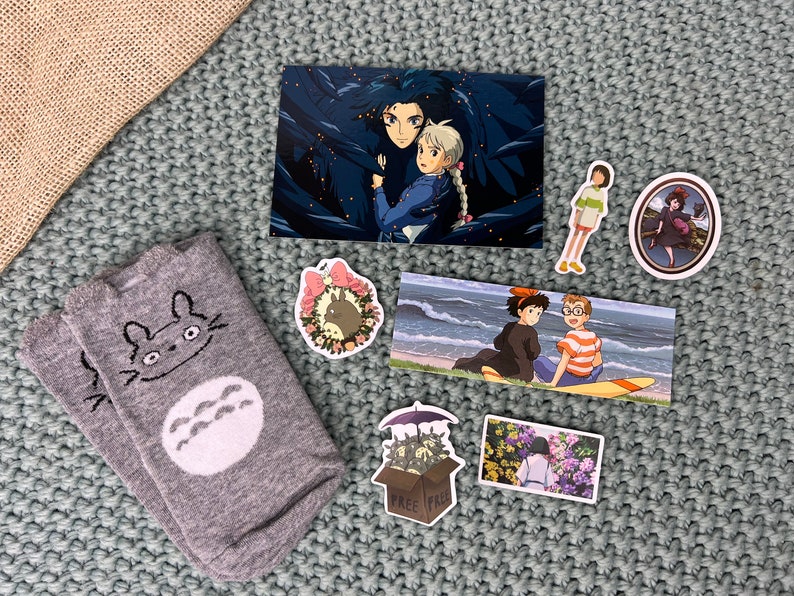 May include: A collection of Studio Ghibli themed items, including a pair of grey socks with a Totoro design, a postcard featuring Howl and Sophie from Howl's Moving Castle, and several stickers featuring characters from different Ghibli films.