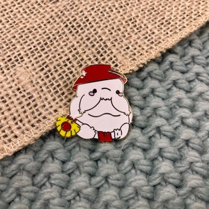 May include: A white enamel pin featuring a cartoon character wearing a red hat and holding a yellow flower. The character has a round body and a small mouth.